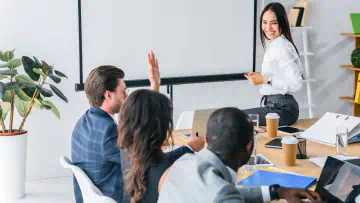 Presenter sits on a conference table. Presentee raises their hand to ask a question.