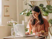 Woman sits at desk and uses laptop. Behind her is a monstera plant