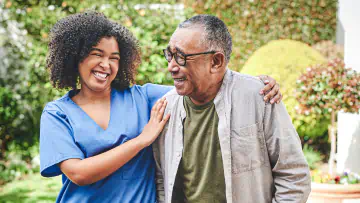 Nurse assists elderly man walking in garden.